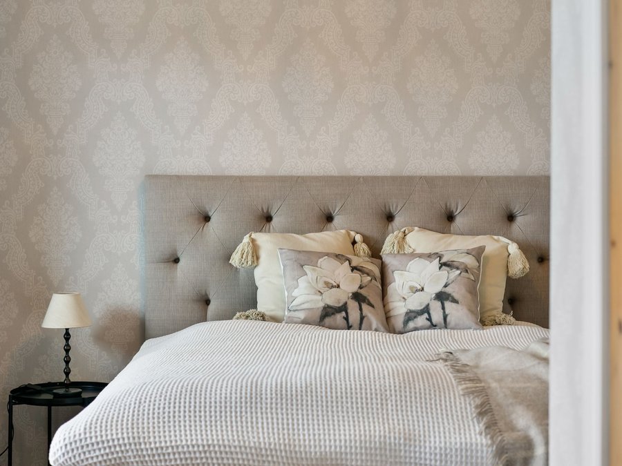 Beige diamond-patterned wallpaper behind a buttoned fabric headboard and bedside lamp