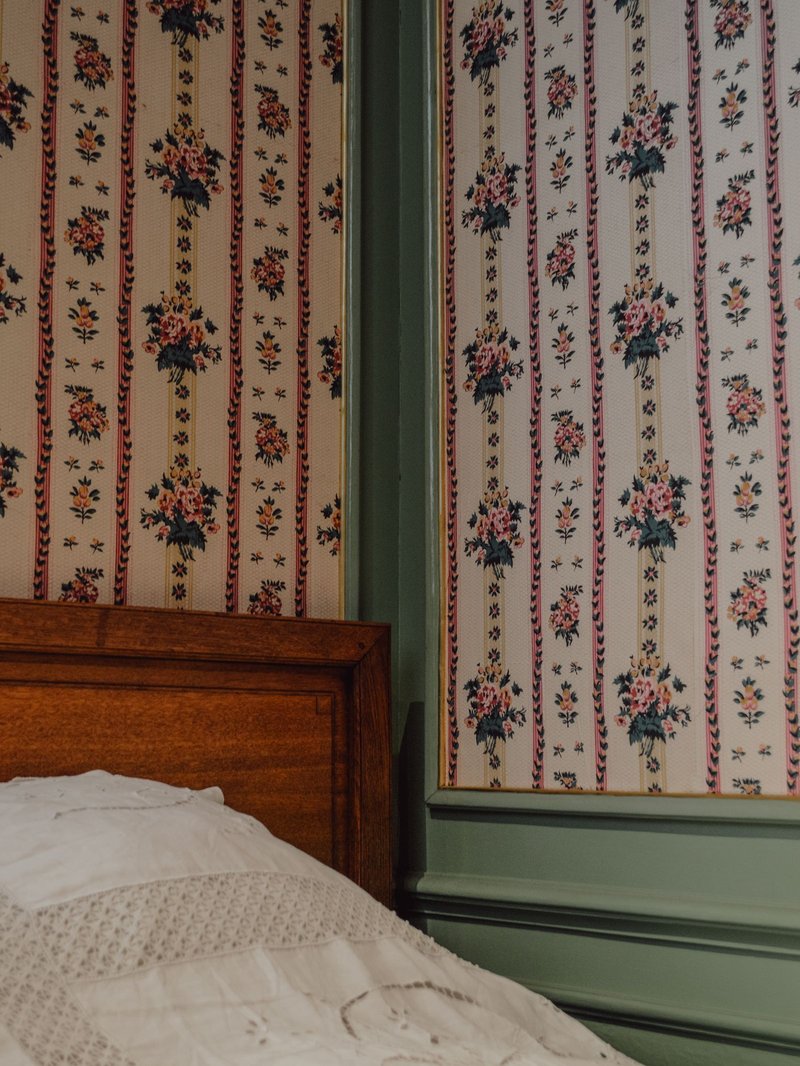 Bedroom corner papered in a pink floral stripe, meeting a sage-painted panel beside a wooden bed