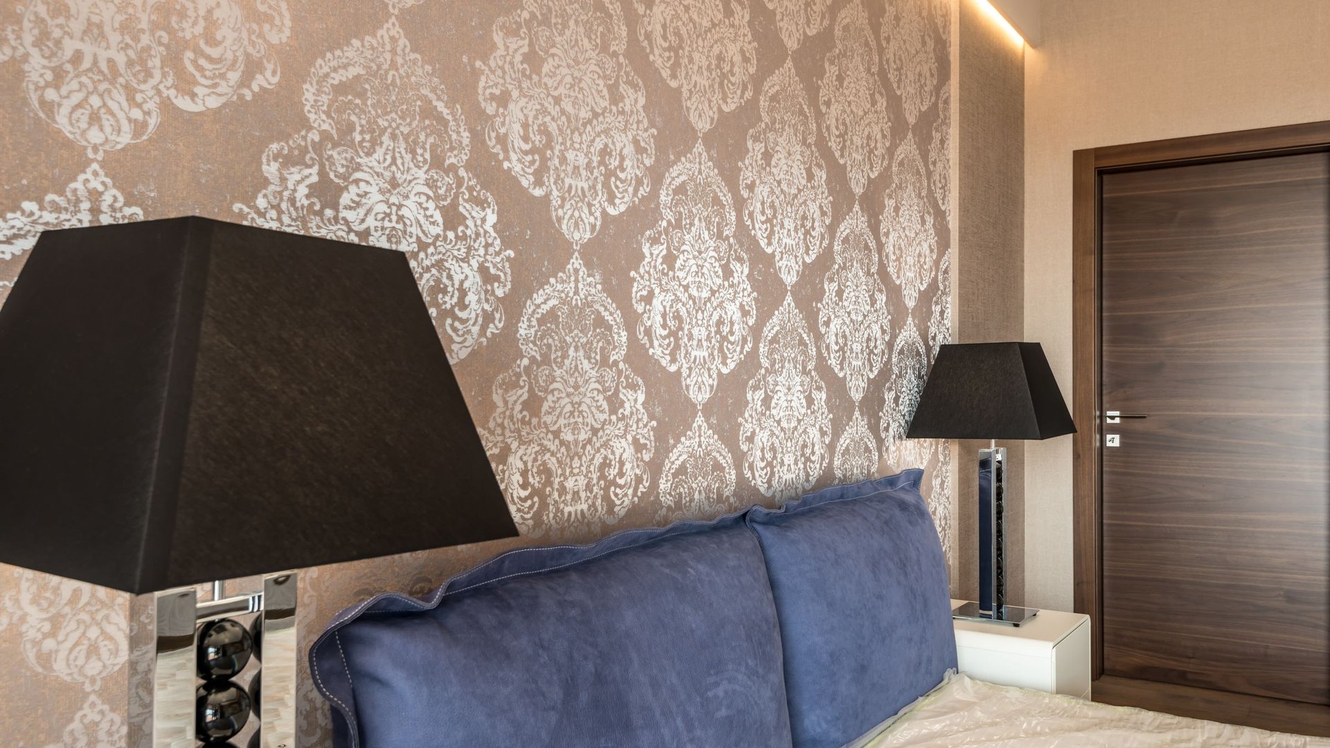 Bedroom wall finished in a soft taupe damask wallpaper, with dark lampshades and a walnut door alongside it